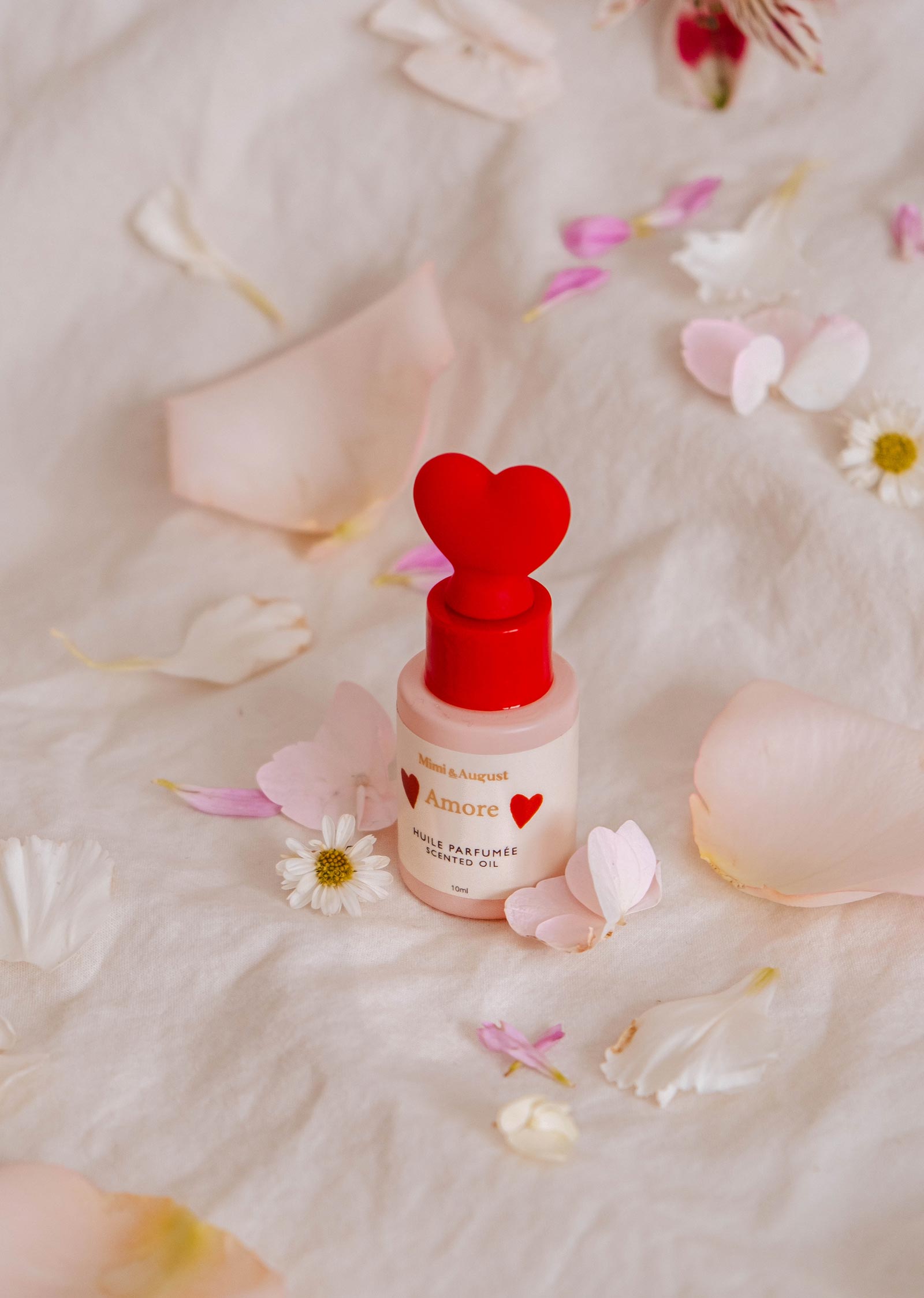 Un petit flacon d'"Huile parfumée - Amore" de Mimi & August, avec un bouchon rouge en forme de cœur, est posé sur un tissu. Son parfum séduisant de pamplemousse et de thé blanc remplit l'air tandis que des pétales de fleurs sont éparpillés autour.