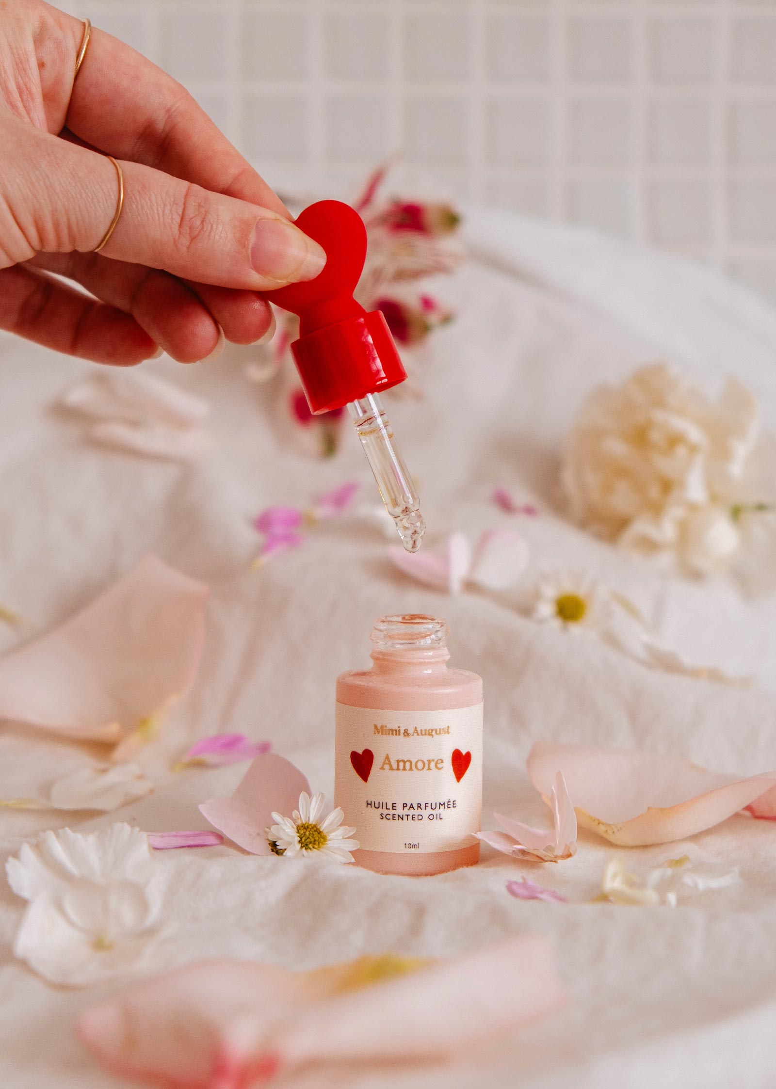 A hand holds a dropper over a small bottle labeled "Scented Oil - Amore" by Mimi & August, surrounded by pink and white flower petals on a soft surface, filling the air with enchanting hints of white tea and grapefruit.