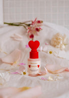 A bottle of Scented Oil - Amore from Mimi & August, featuring a heart-shaped cap, rests among pink and white flower petals. The air is delicately infused with the enchanting fragrance of grapefruit and white tea.