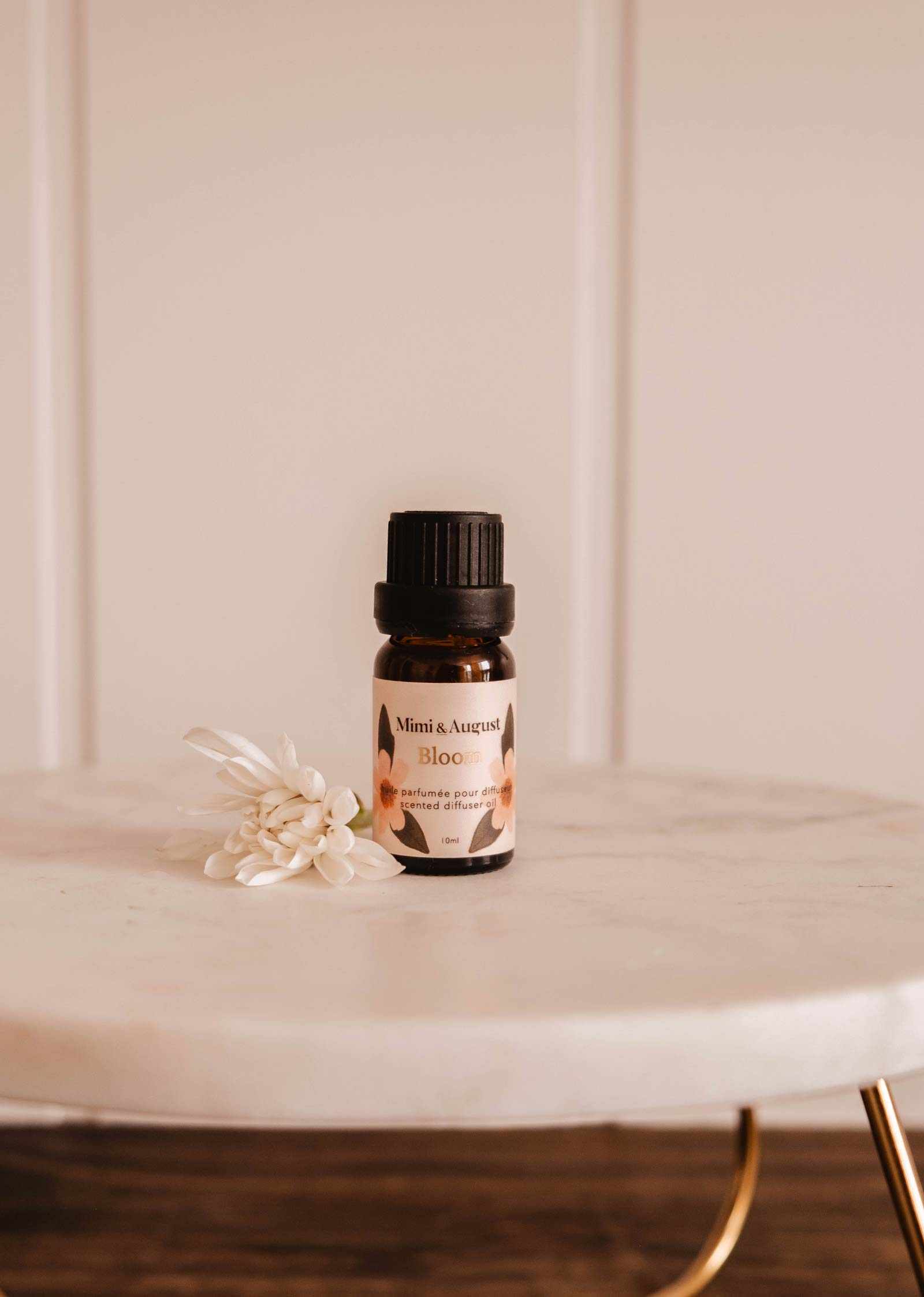 A small Scented Oil - Bloom by Mimi & August rests on a marble surface, releasing a refreshing fragrance with a small white flower beside it.