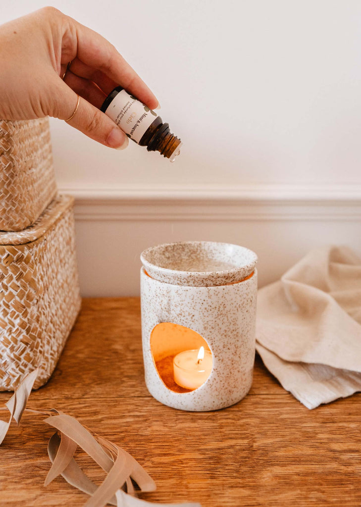 A hand is pouring Mimi & August's Scented Oil - Cielo from a bottle into a ceramic diffuser with a lit tea light candle inside, releasing a calming scent of warm amber. Woven baskets and a beige cloth rest on the wooden surface next to the diffuser.