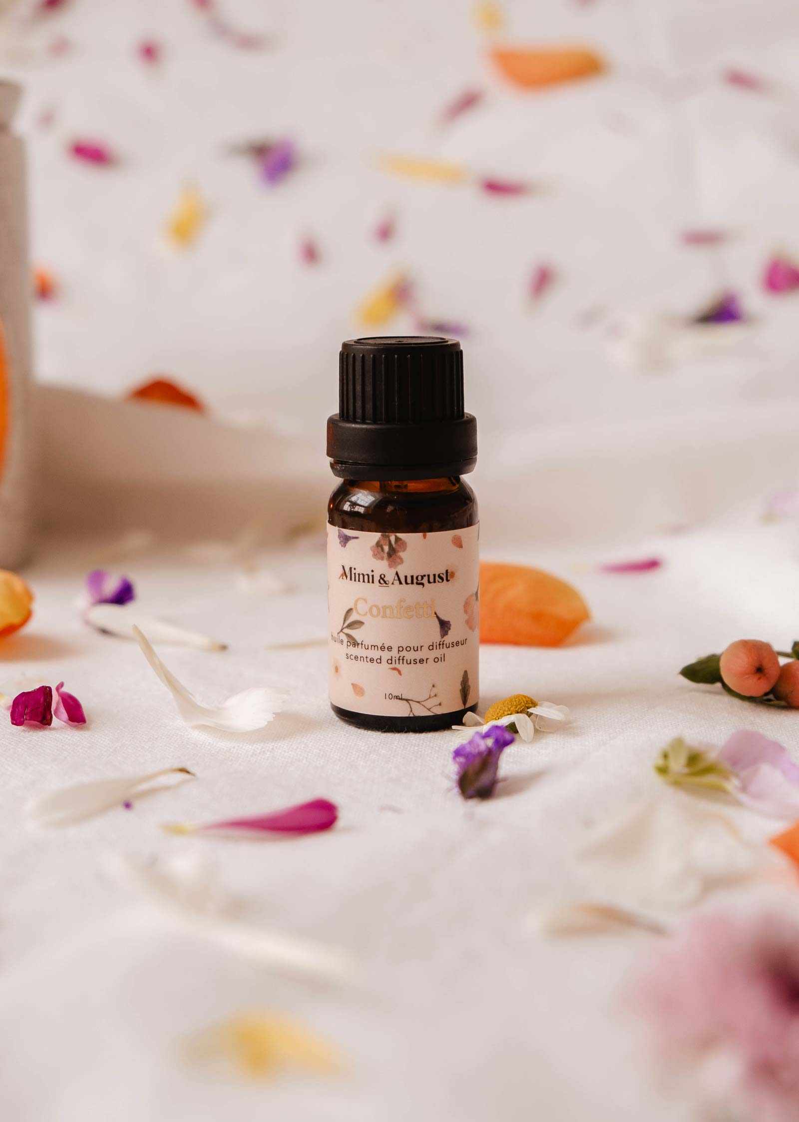 A small amber bottle labeled "Mimi & August Confetti" rests on a white fabric surface, surrounded by scattered colorful flower petals. The Scented Oil - Confetti fills the air with subtle mandarin notes.