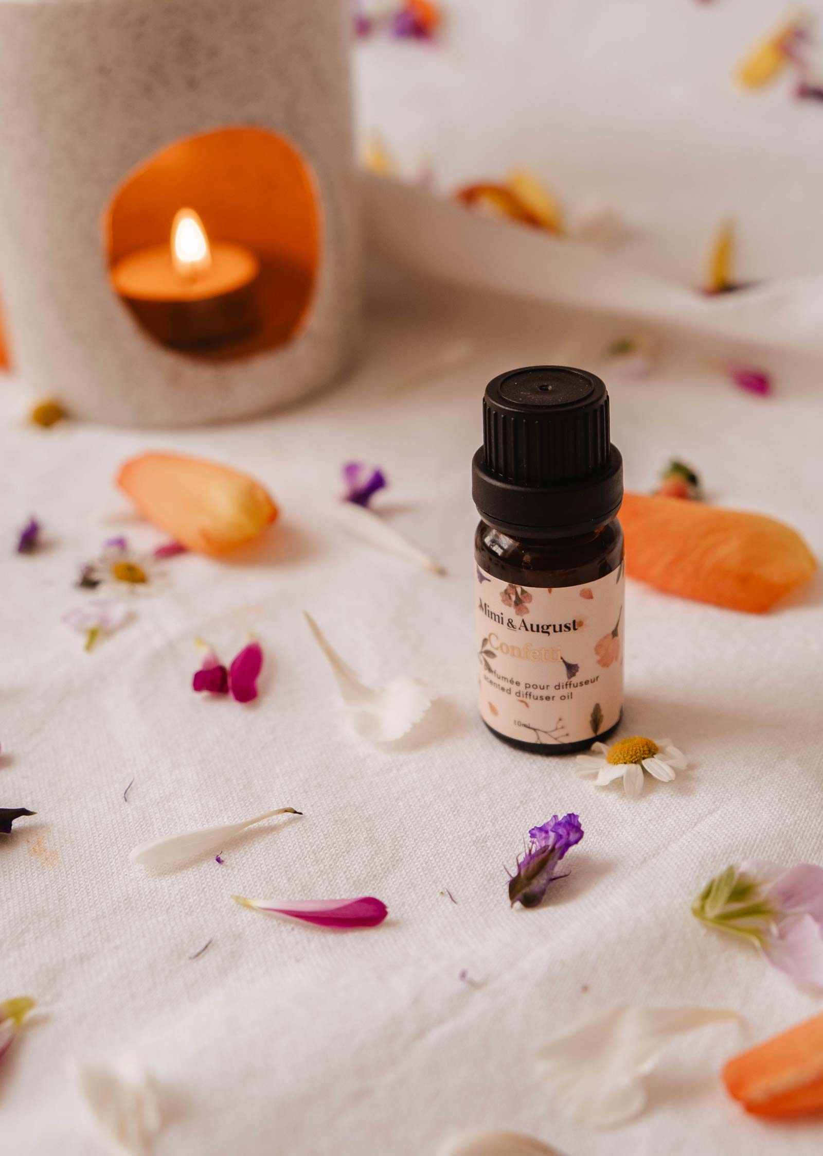 A small bottle of Mimi & August's Scented Oil - Confetti sits on a white cloth with scattered flower petals, releasing its fragrance. A lit candle flickers softly in the background, adding warmth and tranquility to the scene.