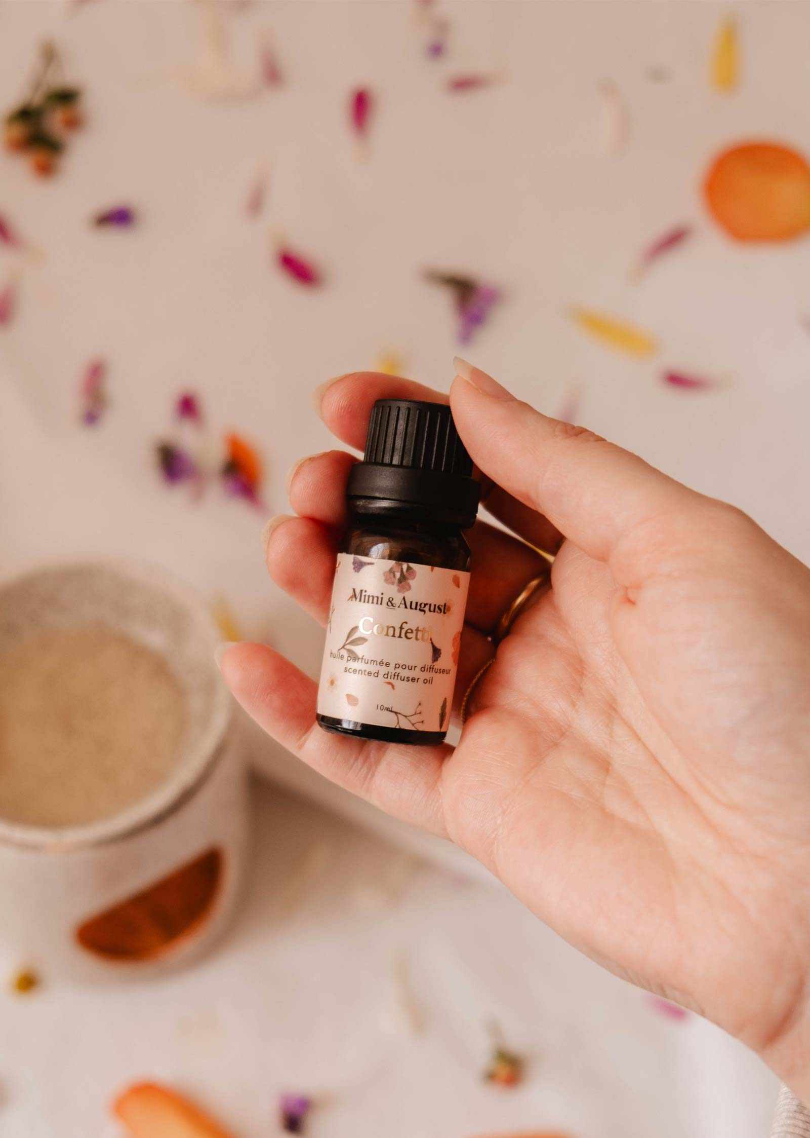 A hand holds a small bottle of Mimi & August's "Scented Oil - Confetti" amidst scattered flower petals and a ceramic oil burner, suggesting a fragrant aroma with hints of mandarin.