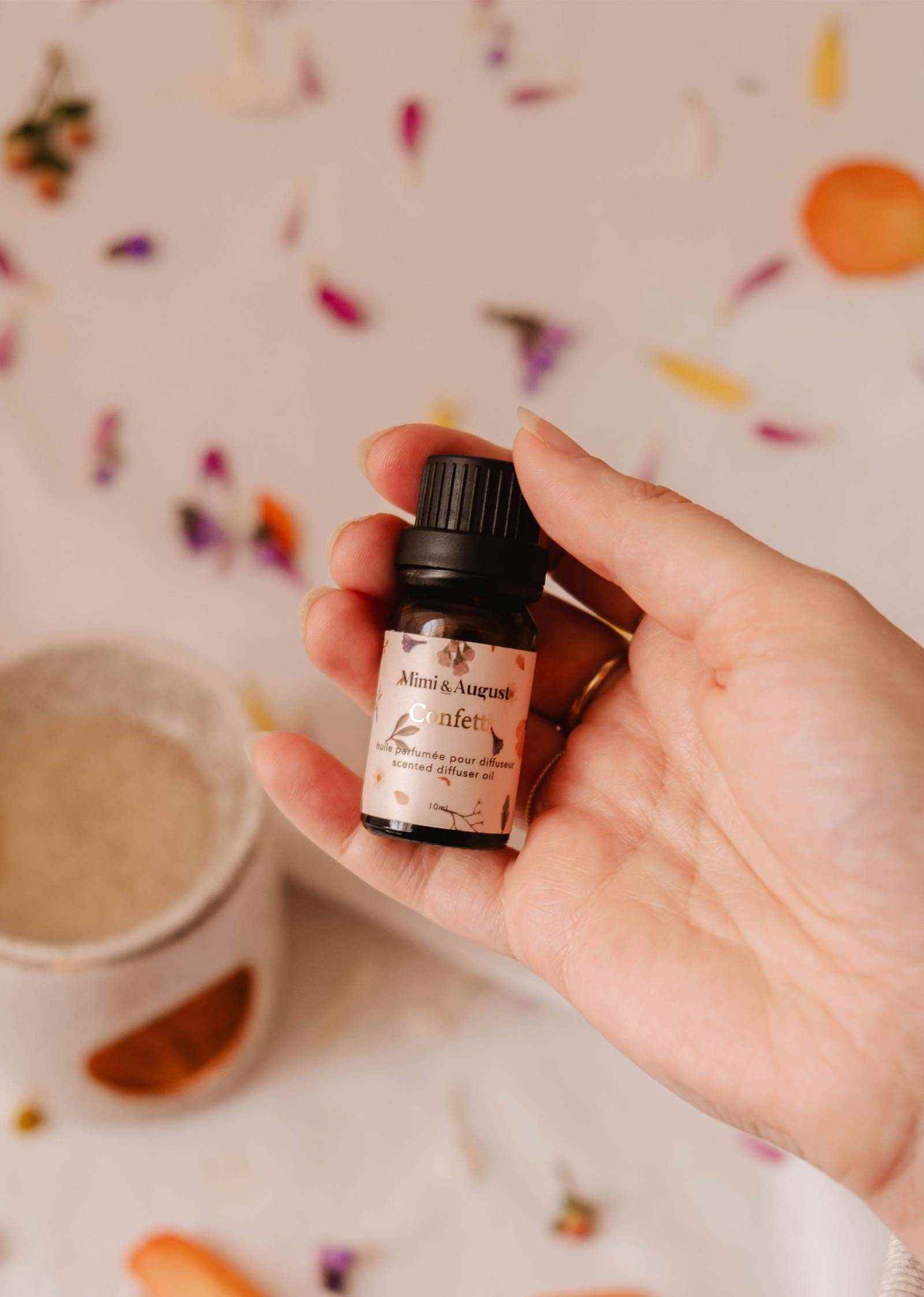A hand holds a small bottle of Mimi & August's "Scented Oil - Confetti" amidst scattered flower petals and a ceramic oil burner, suggesting a fragrant aroma with hints of mandarin.