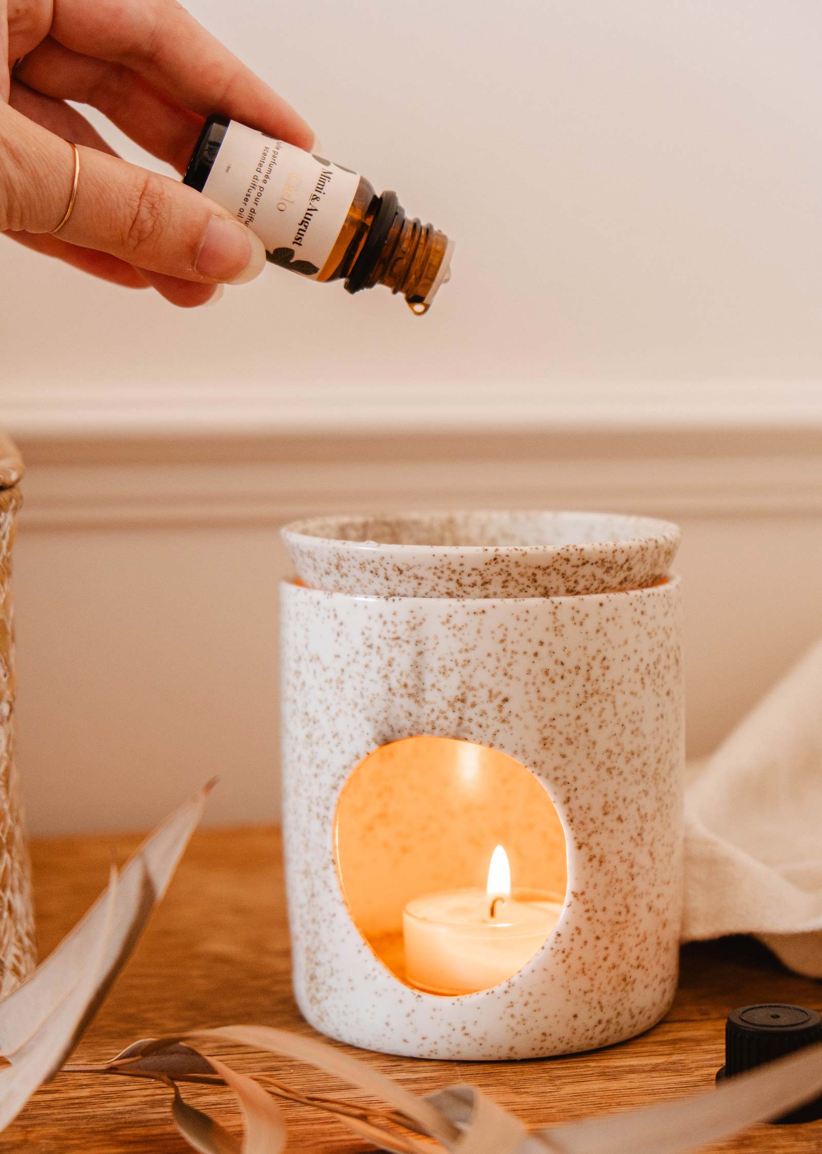 Hand holding a bottle of Mimi & August Scented Oil - Foresta while pouring the forest fragrance into a ceramic diffuser with a lit candle inside, creating a relaxing aroma.