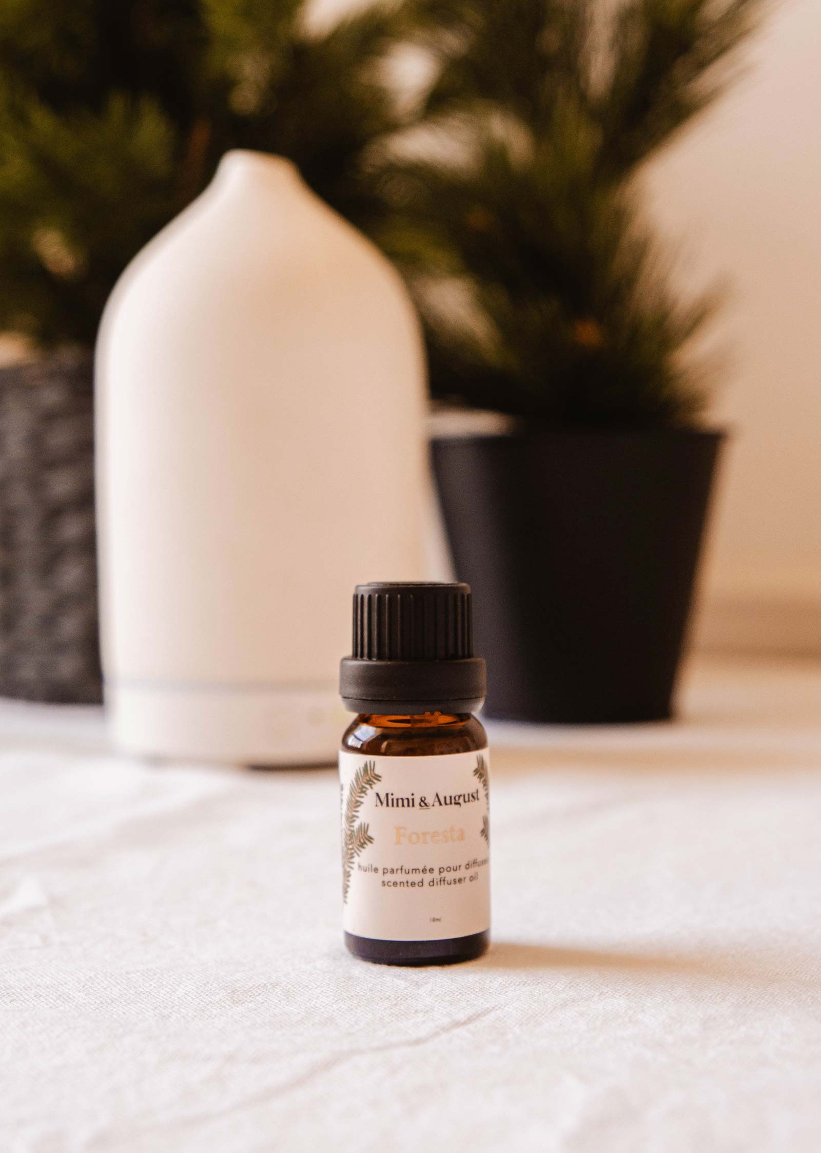 A small brown bottle labeled "Scented Oil - Foresta" from Mimi & August is placed on a white surface, with a white diffuser and a black plant pot in the background. Its fresh forest scent promises to bring a holiday atmosphere into your home.