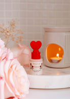A Mimi & August Gift January - Free Scented Oil bottle with a red heart-shaped cap, from the Valentine’s collection, rests on a marble tray beside pink flowers and a lit ceramic candle holder.