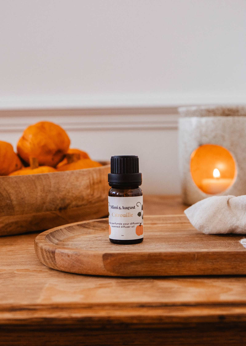 Un petit flacon d'huile parfumée Mimi & August - Citrouille est placé sur un plateau en bois, avec une bougie et un bol d'objets orange en arrière-plan, parfaits pour la relaxation lorsqu'ils sont utilisés dans votre diffuseur.