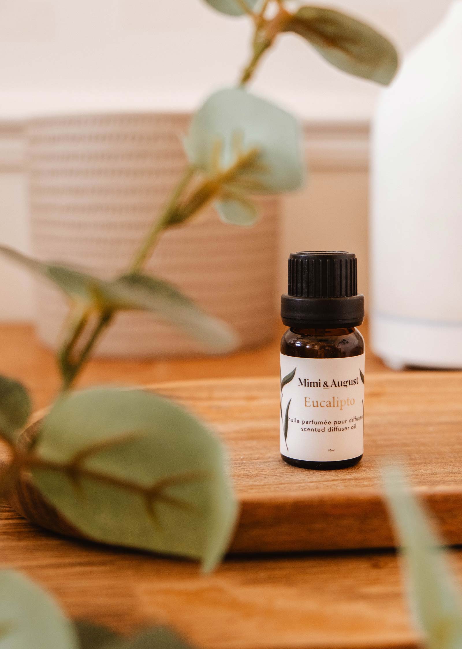 A small bottle labeled "Scented Oil - Eucalipto" from Mimi & August is placed on a wooden surface, surrounded by leaves. The eucalyptus scent offers a relaxing and refreshing atmosphere.