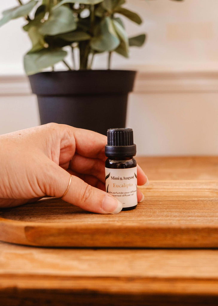 A hand holding a small bottle labeled "Scented Oil - Eucalipto" by Mimi & August on a wooden surface, releasing a soothing eucalyptus scent, with a leafy plant in the background.