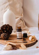 A small bottle of Heirloom Scented Oil by Mimi & August is displayed on a wooden tray adorned with pine cones, dried leaves, and some wood sticks. An aromatherapy diffuser in the background completes the perfect relaxation escape.