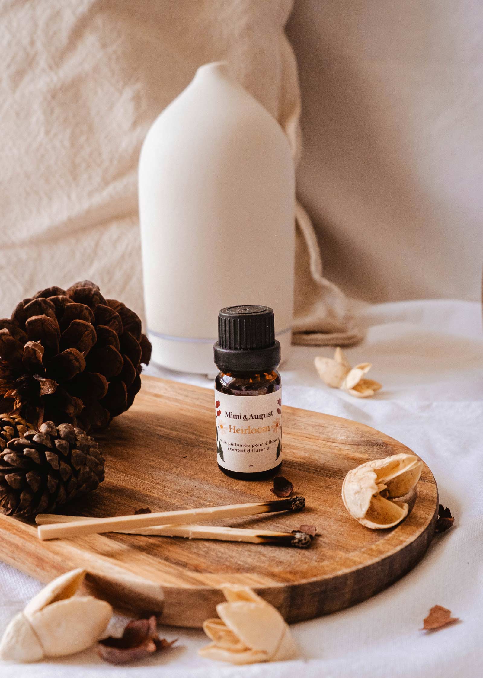 A small bottle of Mimi & August's Scented Oil - Heirloom is placed on a wooden tray next to pinecones, wooden sticks, and a white vase, offering a touch of relaxation and escape.