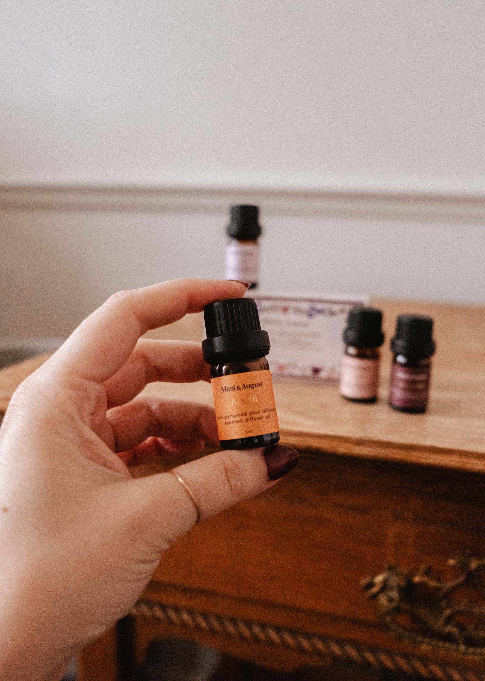 A hand holds a small amber bottle labeled “Scented Oils Discovery Kit” by Mimi & August, with more small bottles and boxes from the collection arranged on a wooden table in the background.