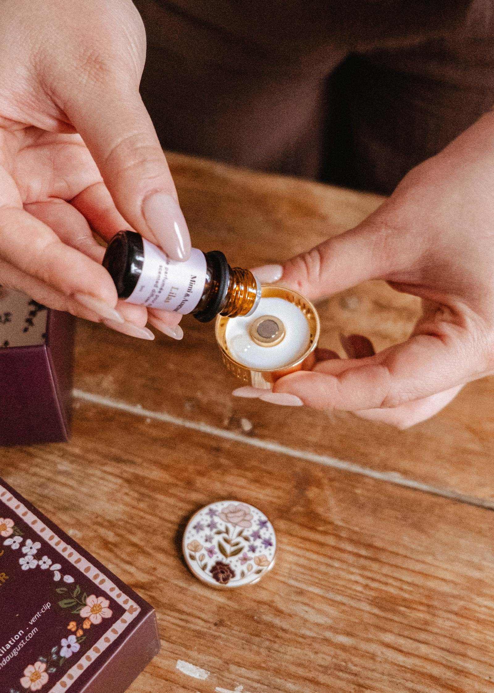 Someone explores their fragrance collection using the Mimi & August Scented Oils Discovery Kit, pouring oils from a small bottle into a round ceramic diffuser with a floral-patterned lid on a wooden table.