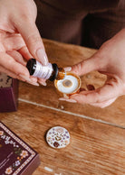 Une personne explore sa collection de fragrances avec le Coffret découverte d'huiles à diffuser de Mimi & August, versant de l'huile d'une petite bouteille dans un diffuseur rond en céramique au couvercle à motifs floraux sur une table en bois.