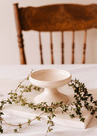 The Mimi & August Serenity Pillar Candle Holder, a handcrafted ceramic piece, sits elegantly on a marble slab with greenery. A wooden chair in the background adds warmth to the serene setting.