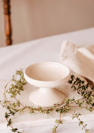 A handcrafted ceramic pedestal bowl rests on a marble surface, surrounded by greenery and a folded beige cloth, exuding the tranquil charm of the Serenity Pillar Candle Holder by Mimi & August.