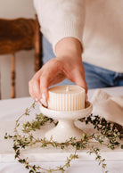 A hand gently places a ribbed candle on the Serenity Pillar Candle Holder by Mimi & August, a handcrafted ceramic piece on a small pedestal, surrounded by lush greenery and complemented by a wooden chair in the background.