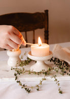 A hand lights a ribbed candle on the Mimi & August Serenity Pillar Candle Holder, which is on a marble stand with green twigs.