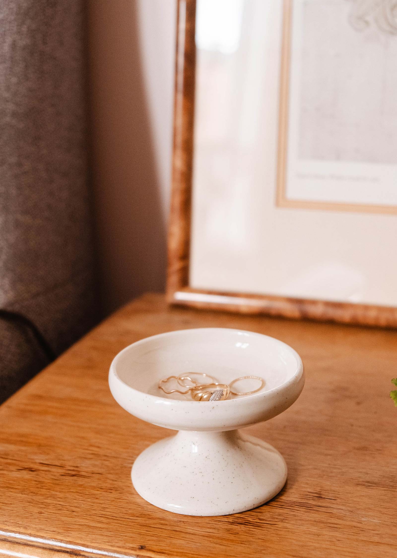 A Serenity Pillar Candle Holder by Mimi & August sits on a wooden surface holding several pieces of jewelry, while a partially visible framed picture enhances the versatile home decor.