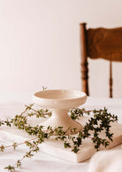 The Serenity Pillar Candle Holder by Mimi & August rests on a marble slab adorned with green vines, next to a wooden chair.