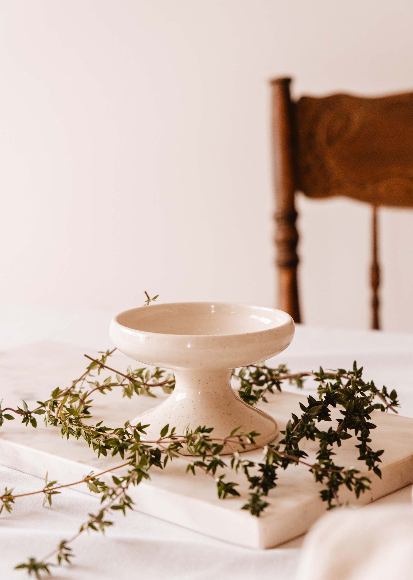 The Serenity Pillar Candle Holder by Mimi & August rests on a marble slab adorned with green vines, next to a wooden chair.