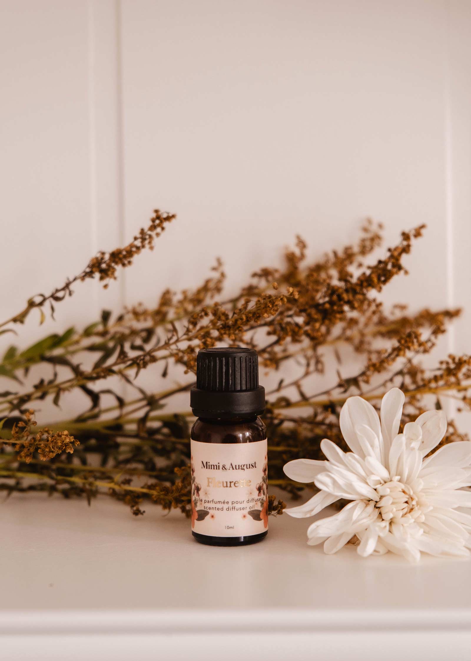 A small bottle with a black cap rests among dried flowers and a full white flower. Its label, "Mimi & August - Set | Exclusive Spring Scented Oil Trio," suggests a spring floral aroma filling the air.