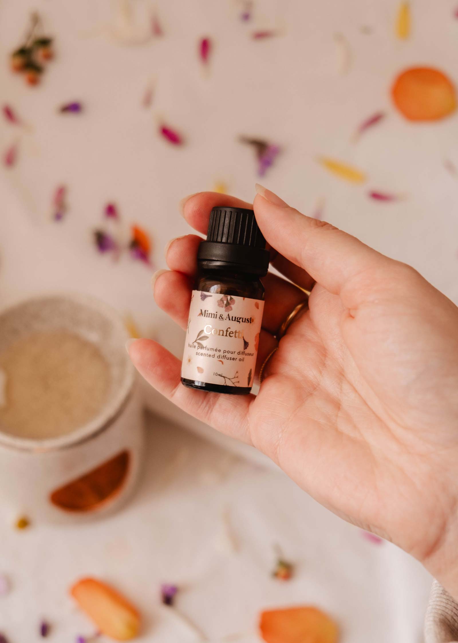 A hand holds a bottle of "Exclusive Spring Scented Oil Trio" by Mimi & August, with a candle holder and flower petals in the background, capturing the essence of floral fragrance.