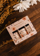 A box of the Mimi & August Set | Exclusive Spring Scented Oil Trio with floral packaging sits on a wooden surface, surrounded by white and brown flowers. Ideal for diffusers to infuse your space with a delightful floral fragrance.