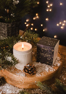 The Shooting Star - Christmas Candle by Mimi & August sits in a ribbed ceramic holder on a wooden tray with faux snow, pine branches, a pine cone, and the candle box, while blurred string lights glow softly in the background.