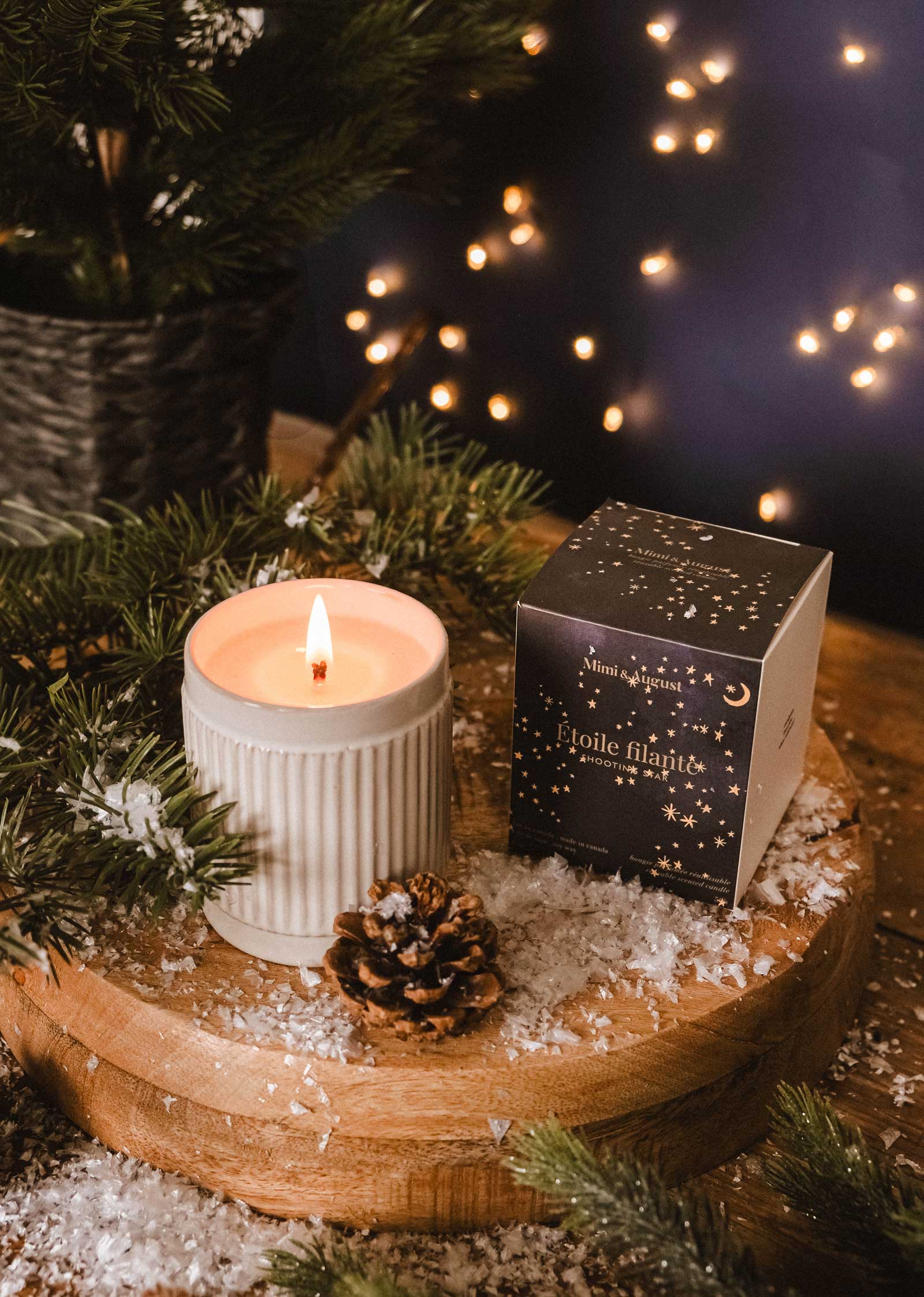 The Shooting Star - Christmas Candle by Mimi & August sits in a ribbed ceramic holder on a wooden tray with faux snow, pine branches, a pine cone, and the candle box, while blurred string lights glow softly in the background.