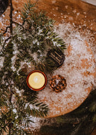 A festive Mimi & August Shooting Star 2 oz candle, pinecone, and fir branches on a wooden snowy surface beside a small “Exotic Forest” label.