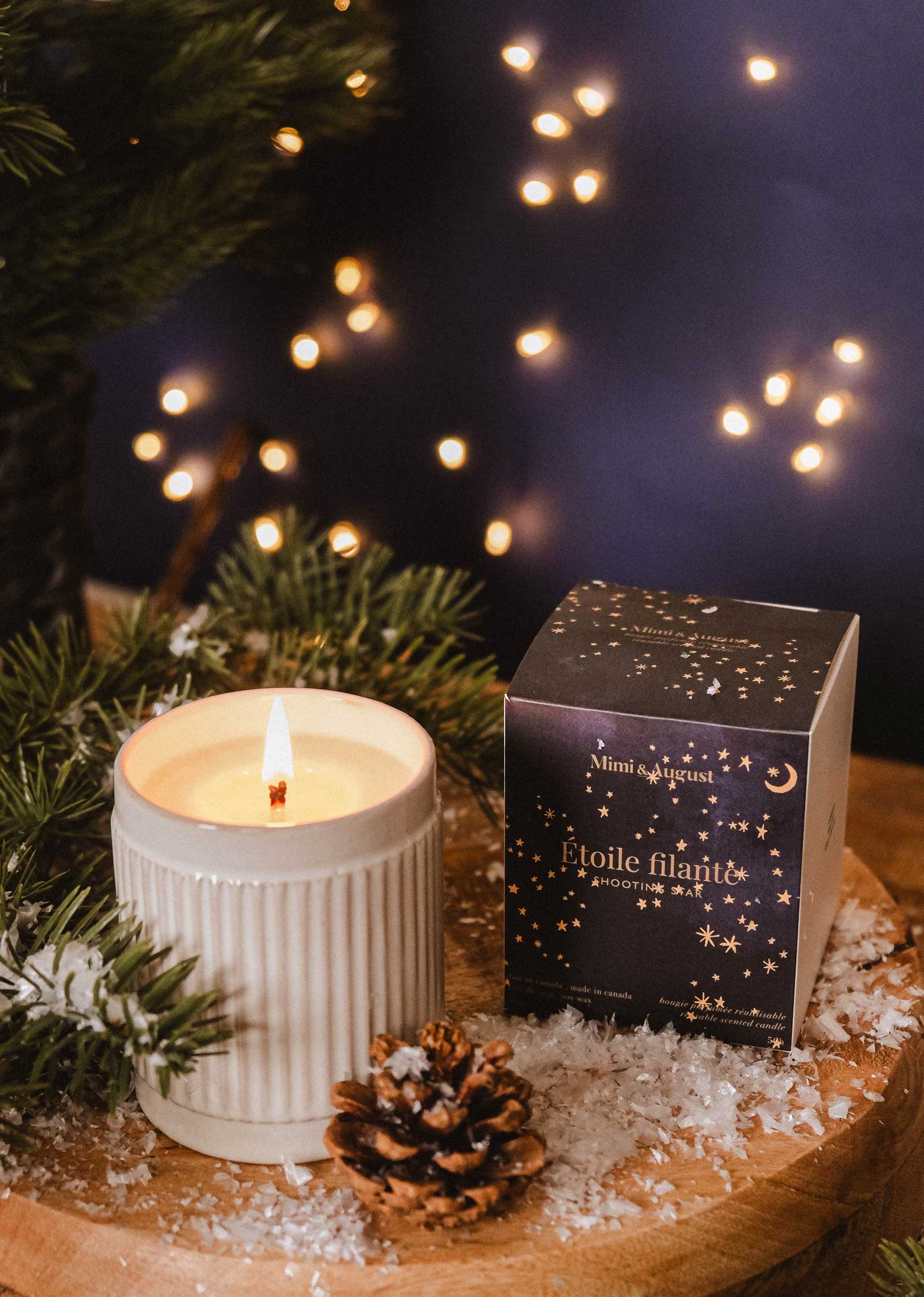 The Shooting Star - Christmas Candle by Mimi & August, in a ribbed container, sits next to a decorated box amid pine branches, a pinecone, and faux snow on wood; blurred lights evoke the cozy spirit of winter nights.