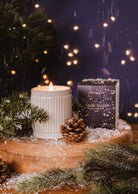 A Shooting Star - Christmas Candle by Mimi & August, a pine cone, and a festive box rest on a wooden surface with faux snow and pine branches, creating cozy holiday vibes with softly glowing lights in the background.