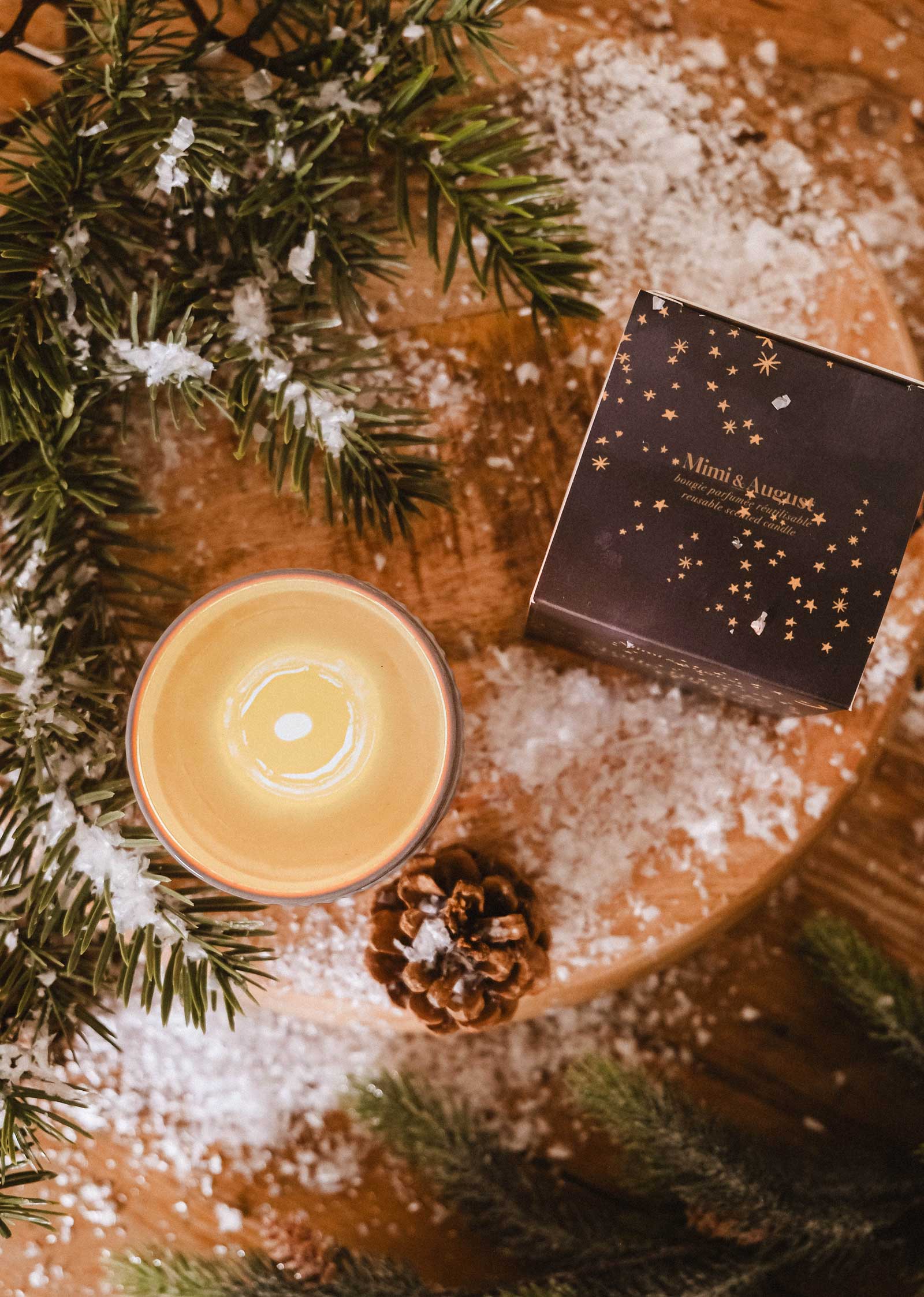 A Mimi & August Shooting Star Christmas Candle, pine cone, artificial snow, pine branches, and a dark starry gift box are arranged on a wooden surface.