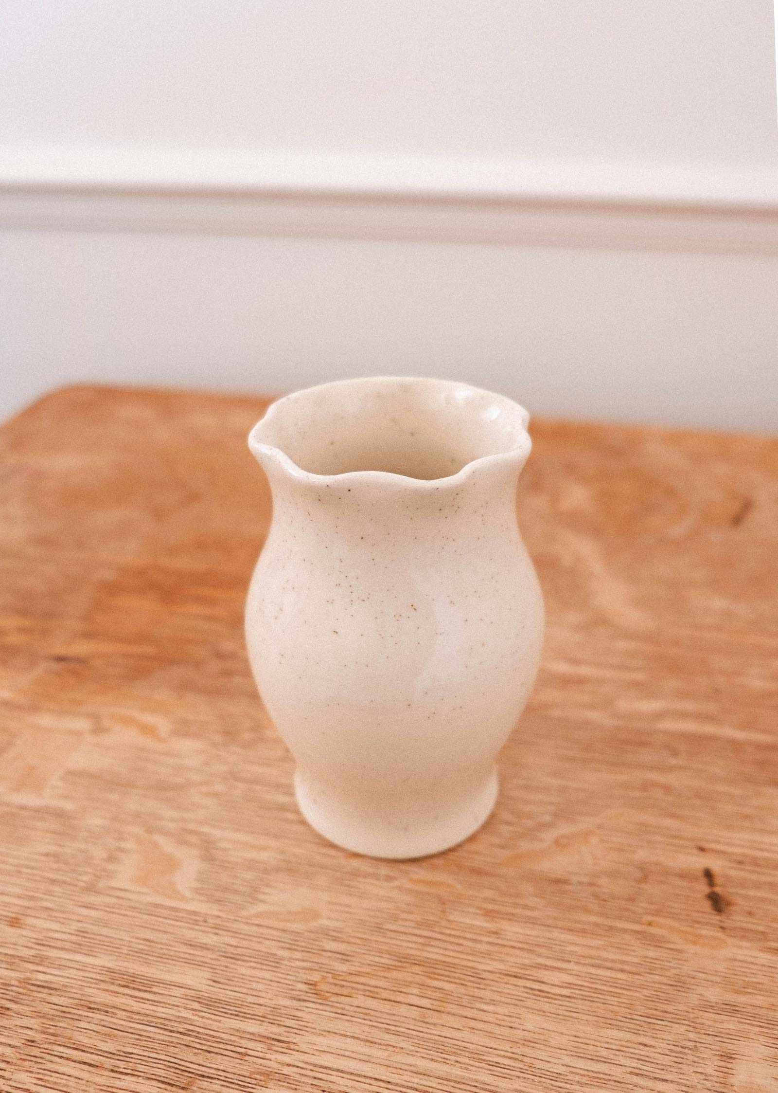 The Silhouette Ceramic Vase by Mimi & August, featuring a cream hue and wavy rim, sits on a wooden surface against a neutral backdrop—a handcrafted piece ideal for enhancing your home décor.