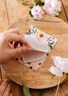 A hand peels a Mimi & August Soft Heart - Sticky Note, shaped like a heart, from a floral notecard on a wooden surface, with pink flowers nearby.