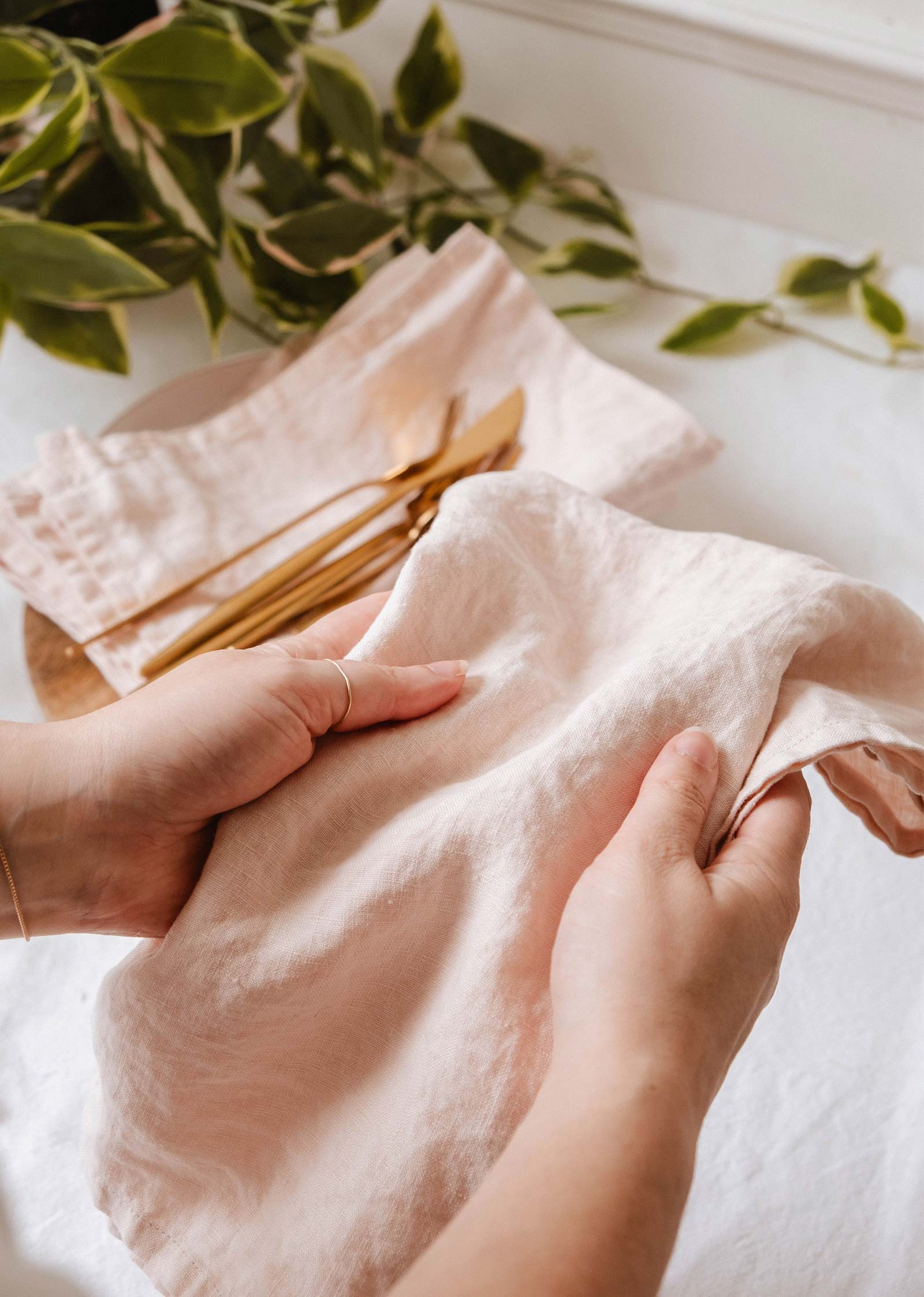 Une personne tient le linge en lin en rose clair, parfait pour faire sécher la vaisselle. D'autres serviettes et des couverts dorés reposent sur une assiette à l'arrière-plan, avec des feuilles vertes à proximité.
