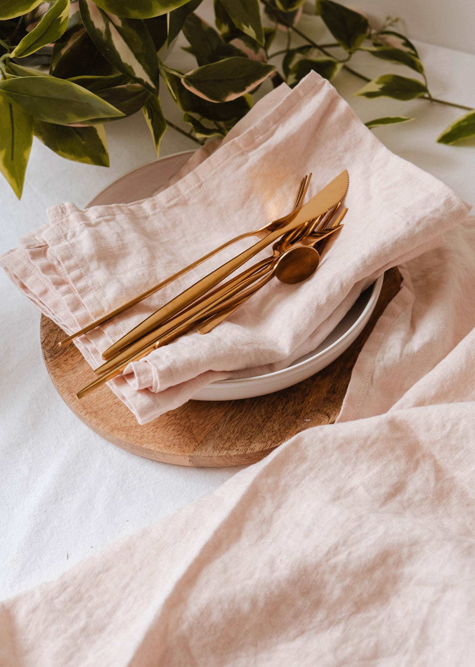 Gold cutlery rests on a white plate and wooden charger, set atop the Mimi & August Tea Towel Soft Linen in pale pink, with leafy greenery in the background for a fresh, airy look.