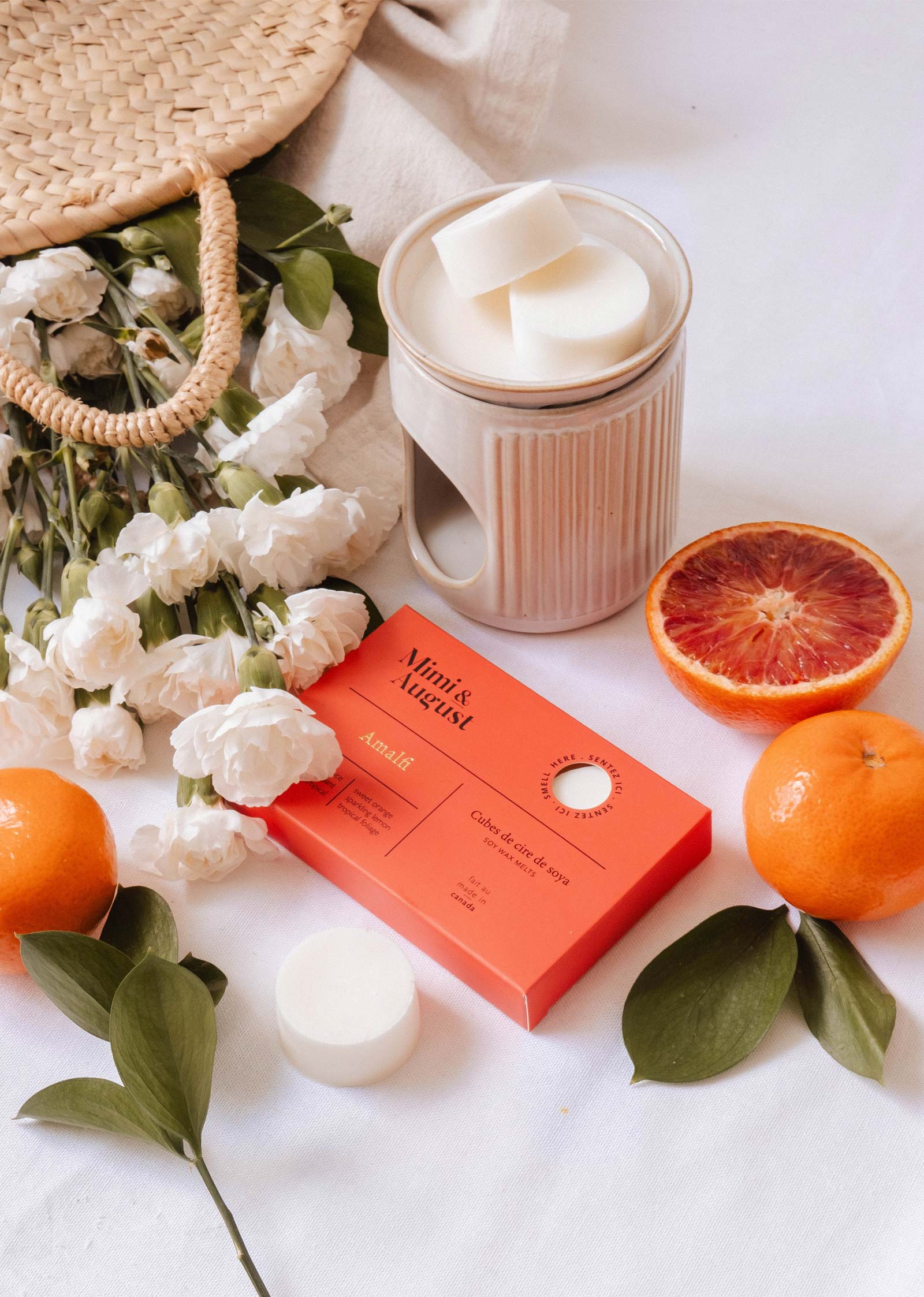 A ceramic mug, red soap box, white flowers, woven bag, orange, tangerine, and sliced blood orange on a white surface reflect the lasting scent of Mimi & August’s Soy Wax Melts - Amalfi made with natural soy wax.
