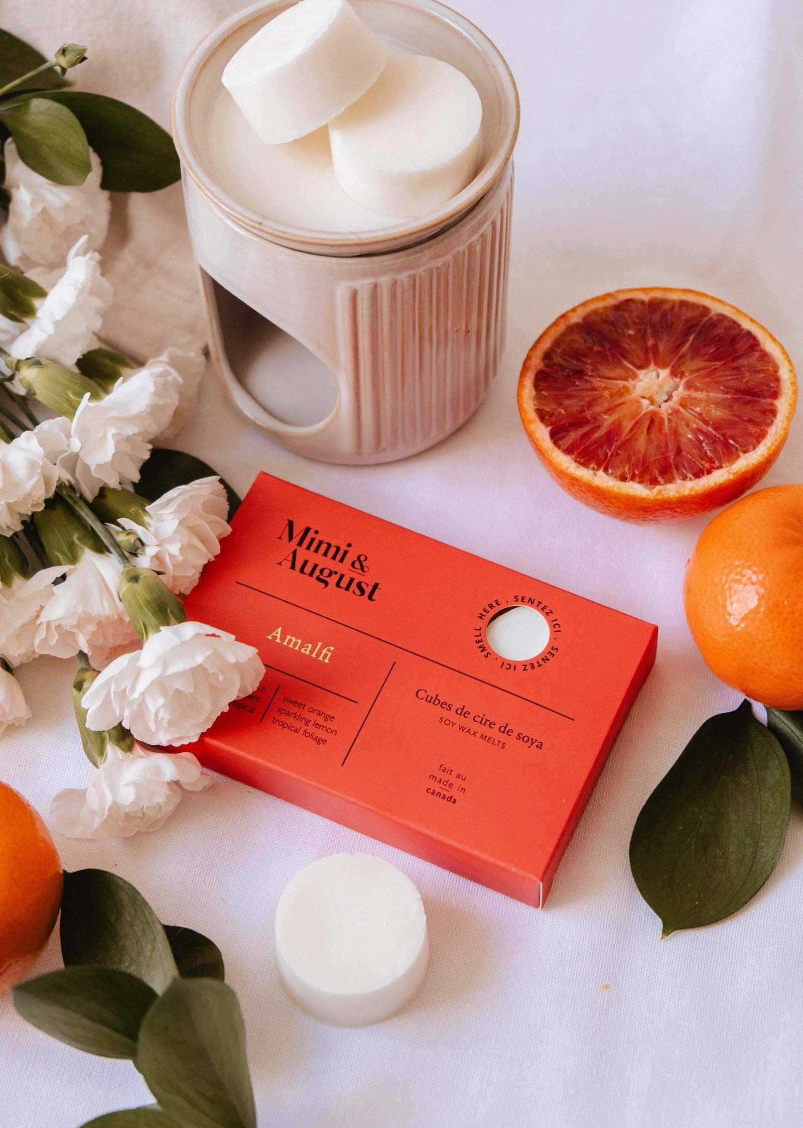 The Mimi & August Soy Wax Melts - Amalfi set offers a long-lasting fragrance and comes with a red gift box, ceramic wax warmer, white flowers, green leaves, an orange, and a halved blood orange on a white surface.