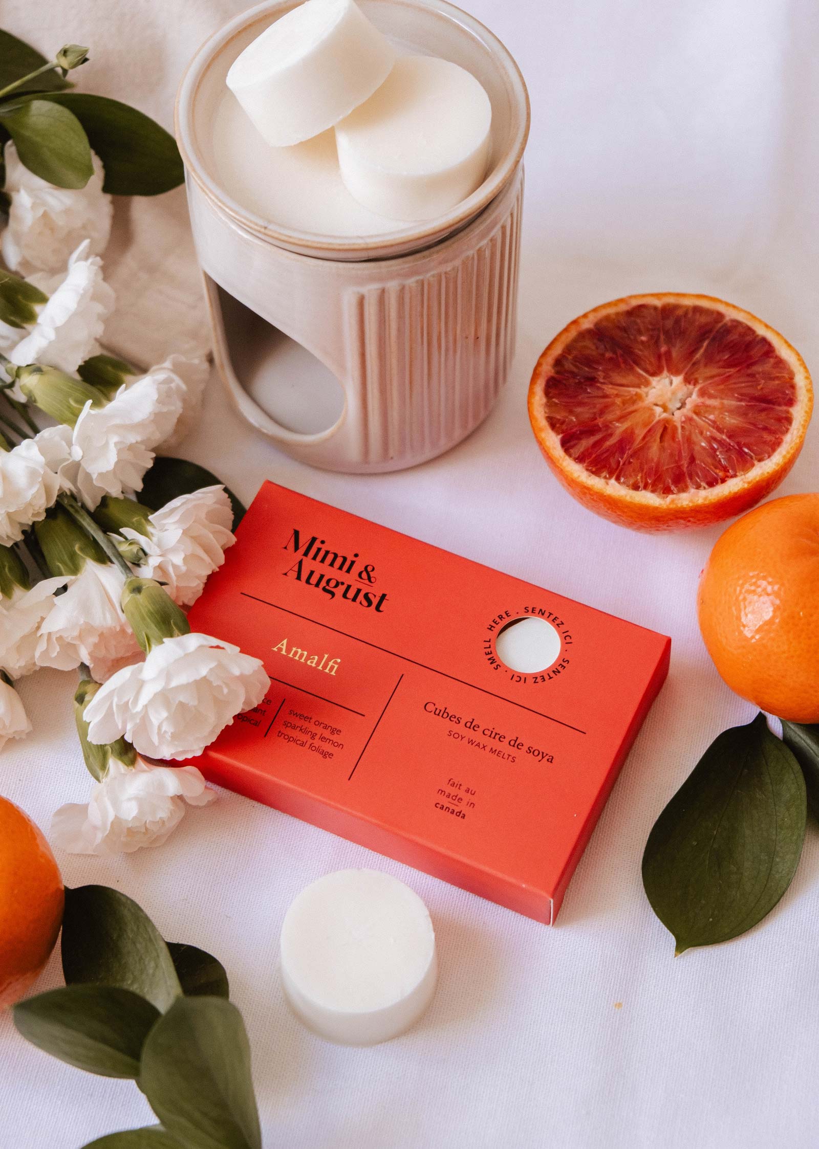 The Mimi & August Soy Wax Melts - Amalfi set offers a long-lasting fragrance and comes with a red gift box, ceramic wax warmer, white flowers, green leaves, an orange, and a halved blood orange on a white surface.