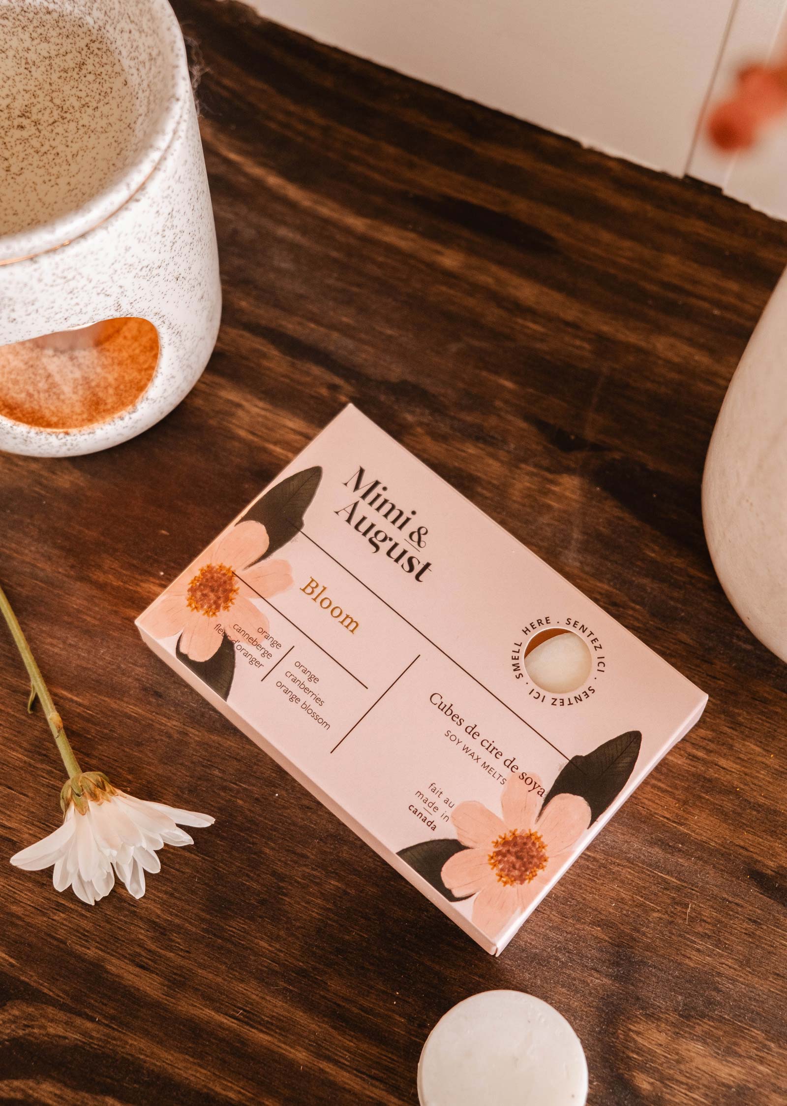 A box of "Mimi & August" Soy Wax Melts - Bloom sits on a wooden surface, surrounded by ceramic cups and a small white flower, exuding an eco-friendly charm akin to a solid face cleanser.