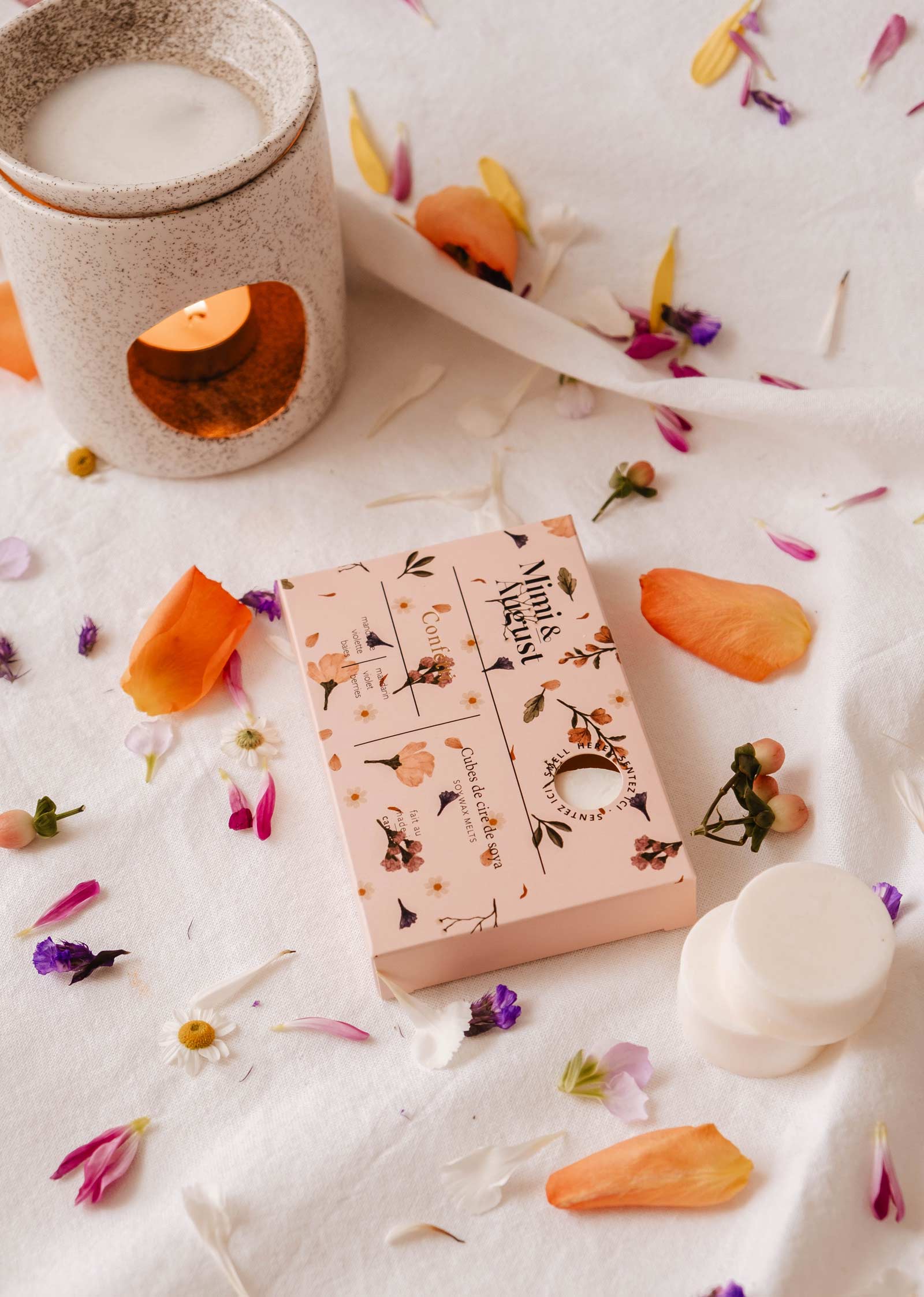 Un diffuseur et une boîte des cubes de cire à motifs floraux reposent sur un tissu blanc, entourés de pétales de fleurs, libérant le parfum écologique et durable des cires de soja fondues Mimi & August - Confetti.