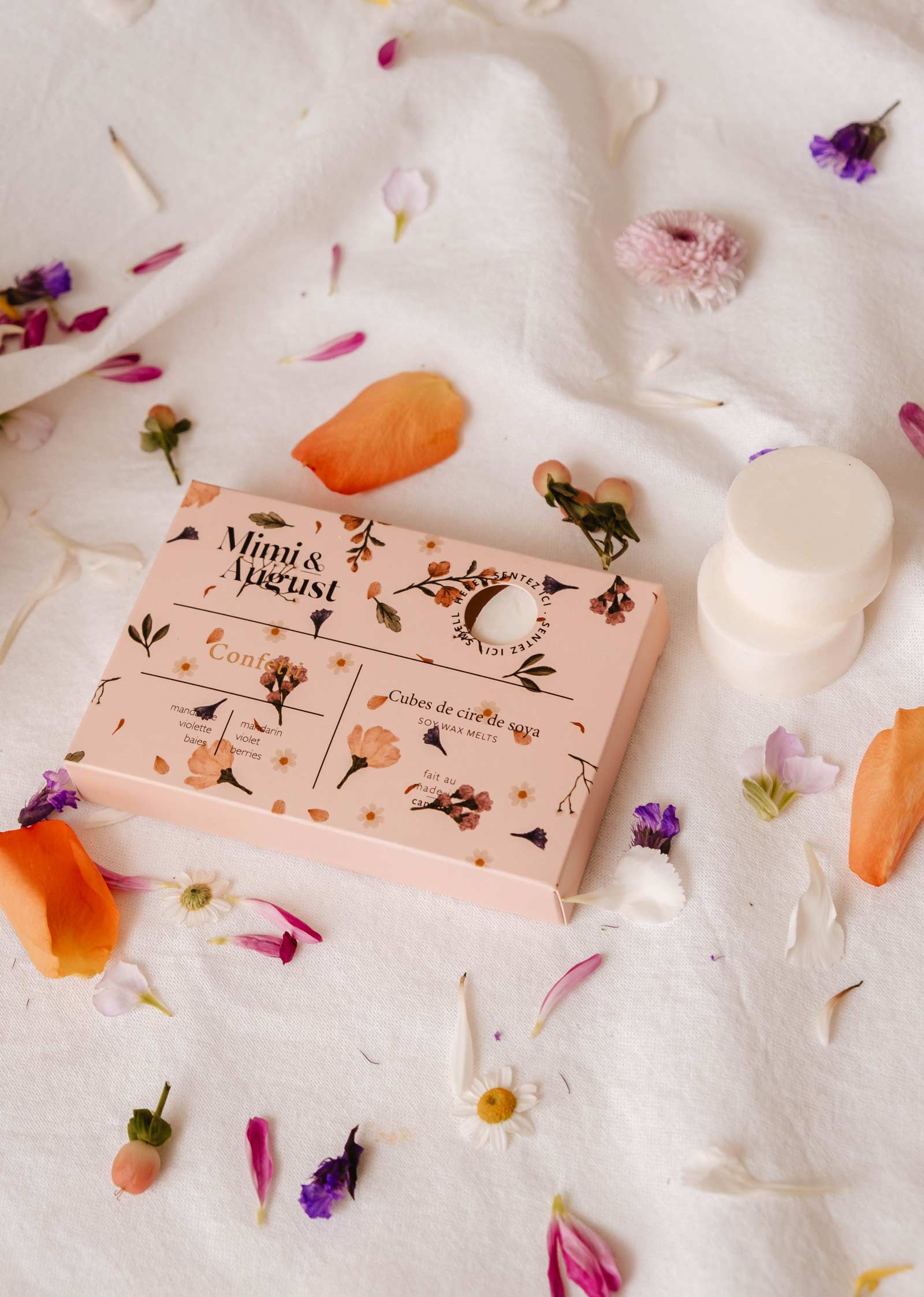 A box labeled "Mimi & August" containing Soy Wax Melts - Confetti, celebrated for their eco-friendly design and long-lasting fragrance, rests on white fabric surrounded by scattered flower petals.