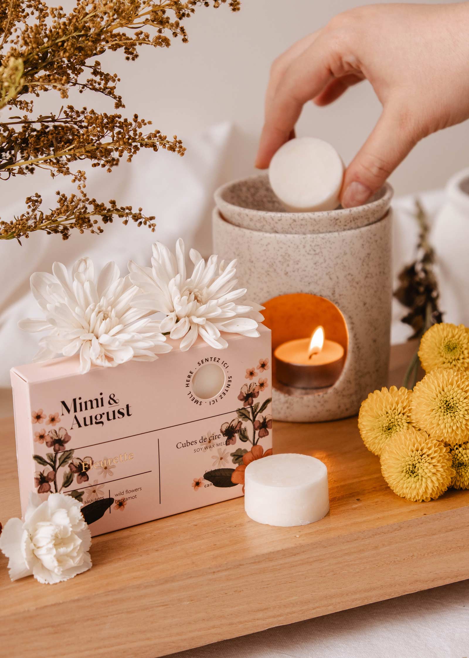 Une main place un cube de cire de soja Fleurette dans un diffuseur avec une bougie allumée. À côté, une boîte rose Mimi & August à motif floral est entourée de fleurs blanches et jaunes sur du bois, promettant un parfum de longue durée.