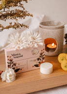 À côté d'une boîte de cubes de cire Mimi & August, ornée de fleurs blanches, une bougie parfumée fabriquée à partir de cire de soja, respectueuse de l'environnement, brûle à côté d'une délicate composition florale sur une surface en bois.