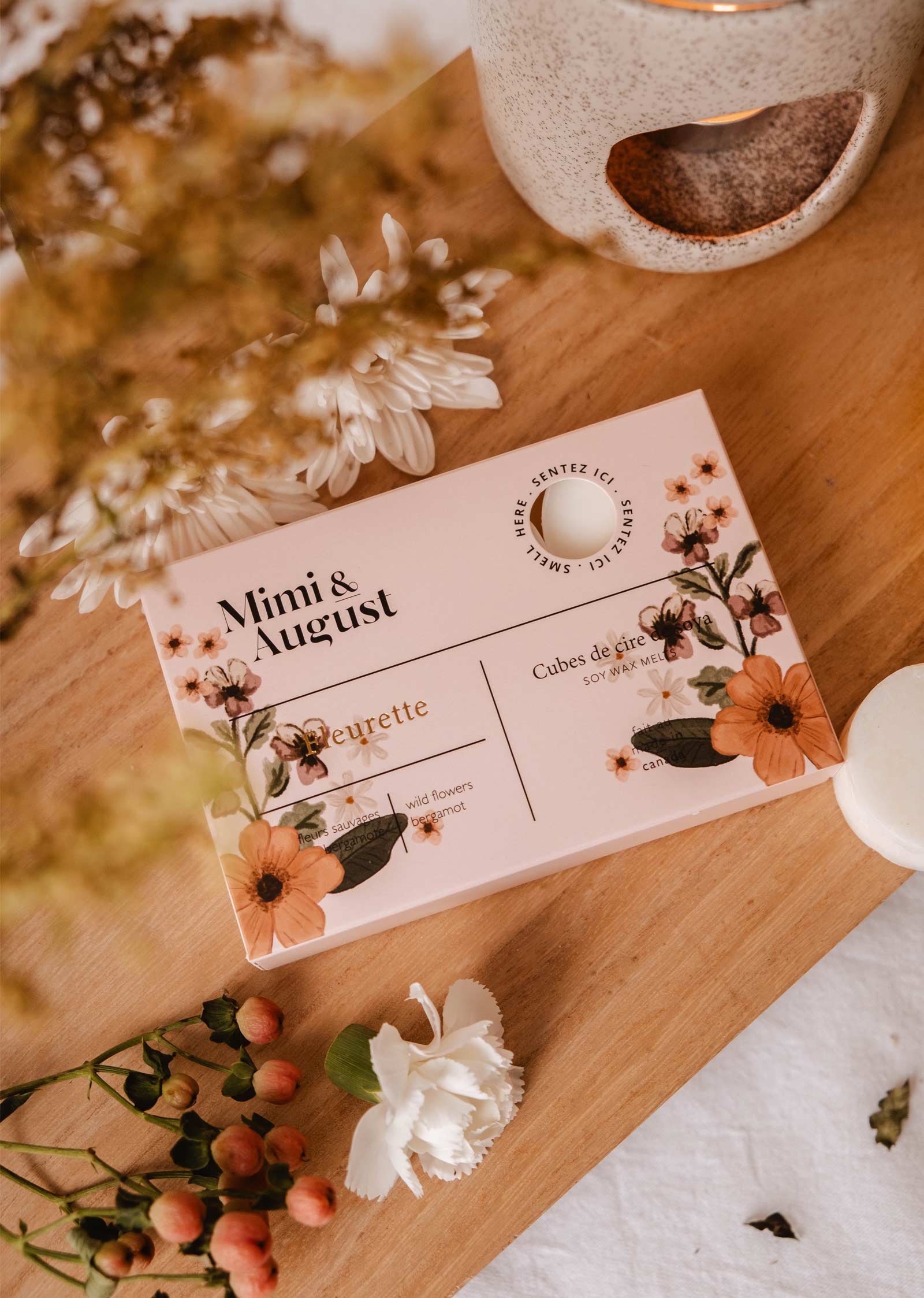 A floral-printed box of Mimi & August's Fleurette Soy Wax Melts sits on wood with a lit wax warmer, surrounded by white flowers and berries, creating an eco-friendly ambiance and long-lasting fragrance.