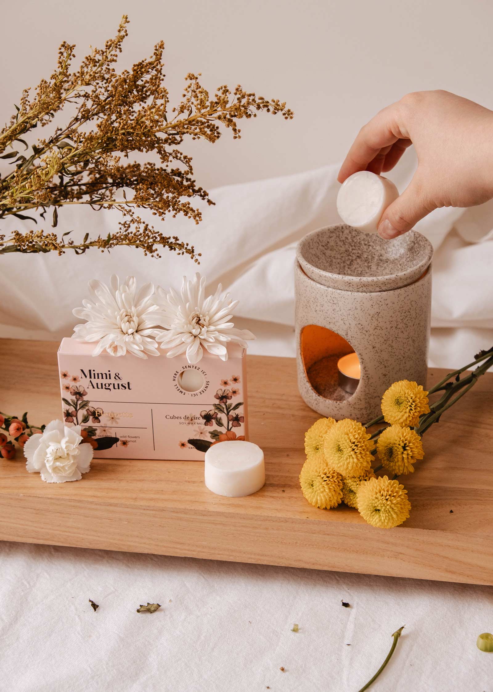 A hand gently places a Soy Wax Melts - Fleurette by Mimi & August in a ceramic warmer on a wooden tray, surrounded by flowers and a box labeled "Mimi & August," promising an eco-friendly and long-lasting fragrance.